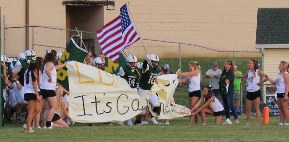 The football team running through the banner on game night! One player is carrying the American flag.